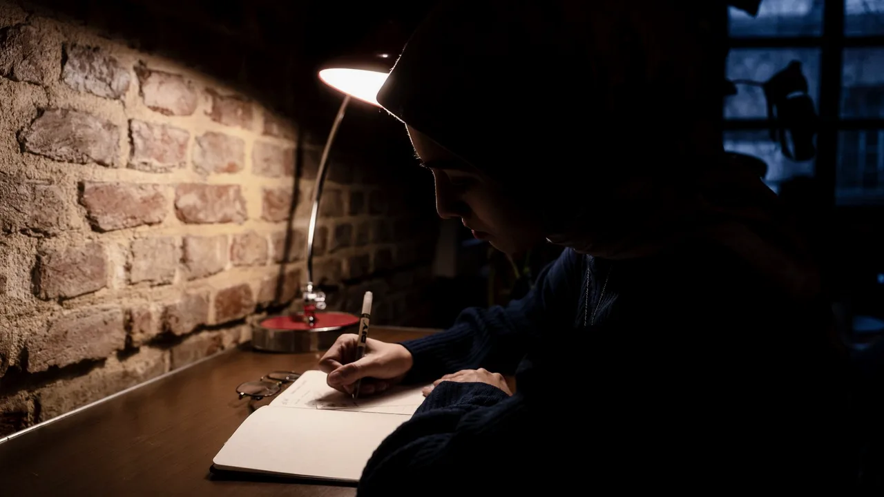 Pessoa escrevendo num caderno sob a luz de uma luminária de mesa, em ambiente escuro à noite