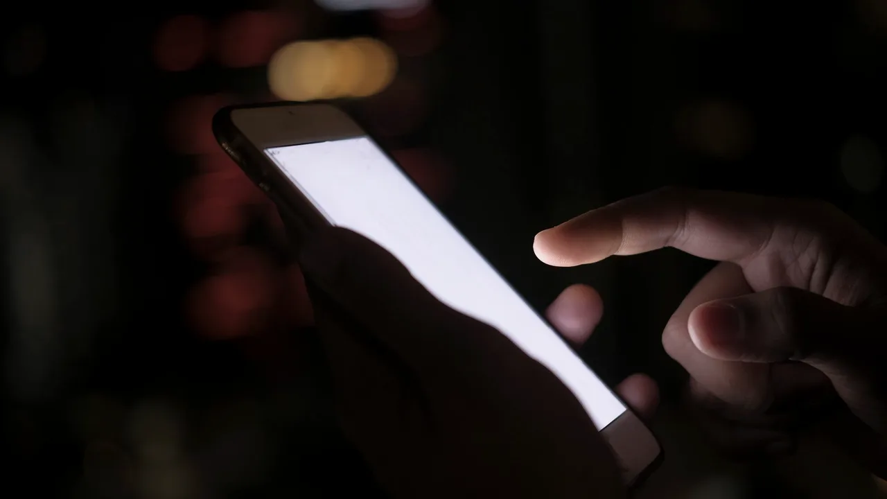 Mão tocando a tela iluminada de um celular no escuro, com luzes desfocadas em vermelho e dourado ao fundo