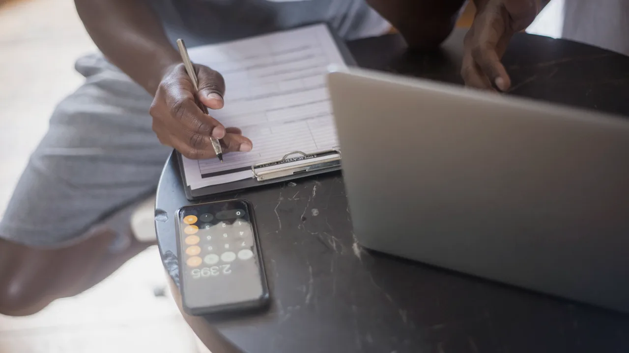 pessoa anotando itens numa prancheta ao lado de um notebook aberto sobre mesa escura, com o celular ao lado