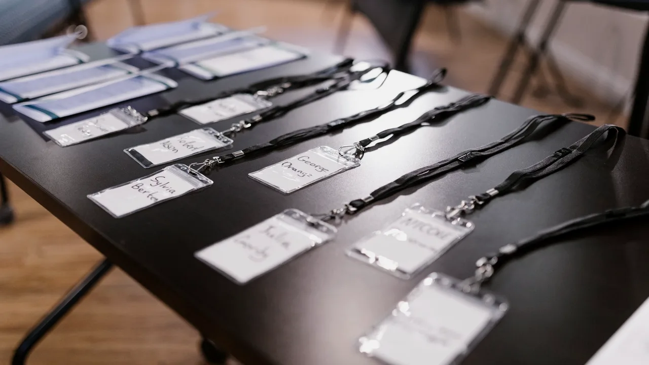 Crachás de identificação com nomes escritos à mão, presos a cordões pretos, alinhados sobre uma mesa escura