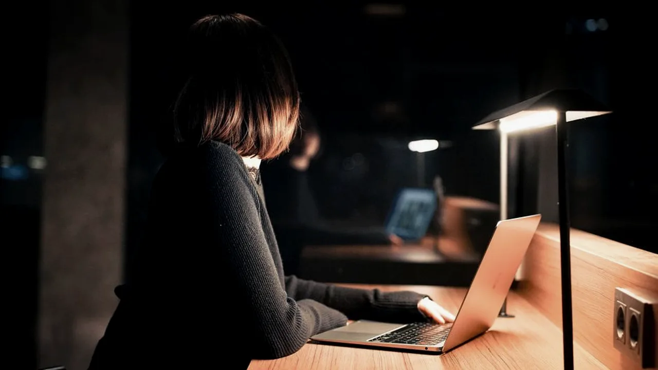 Mulher trabalhando no laptop num ambiente escuro, iluminada pela luz quente de um abajur sobre a mesa