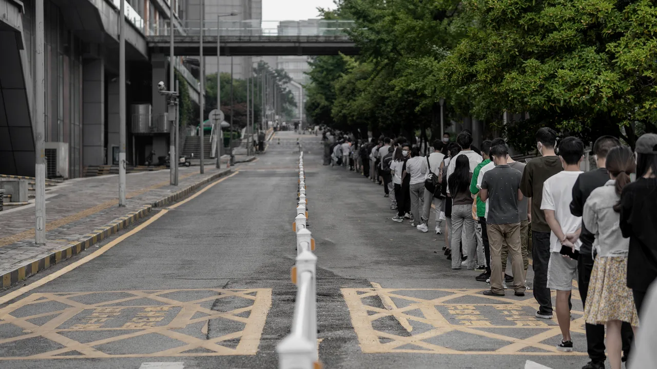 Fila enorme de pessoas em pé na beira de uma rua, uma atrás da outra, sumindo ao longe