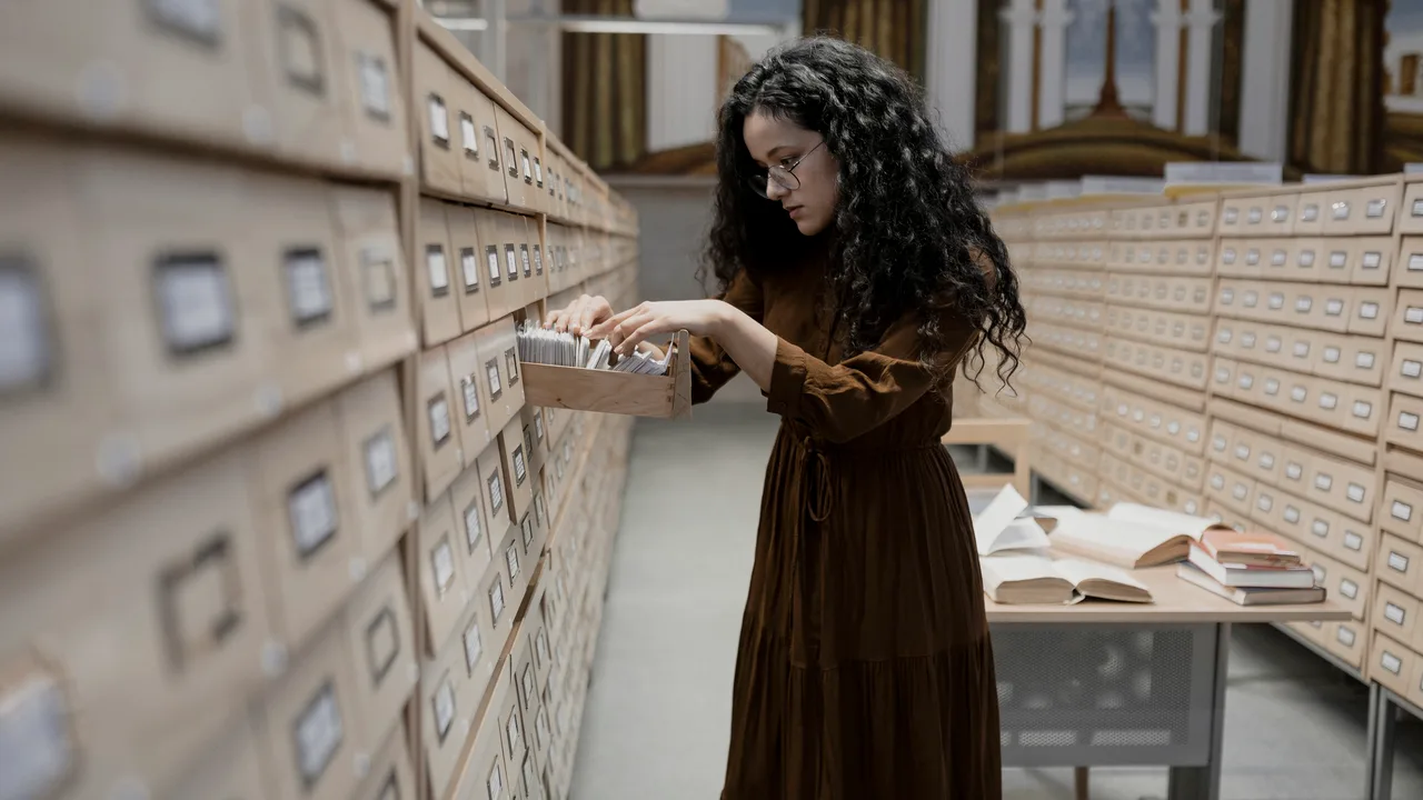 Mulher abrindo a gaveta de um fichário de arquivo, diante de uma parede de gavetas com fichas catalogadas