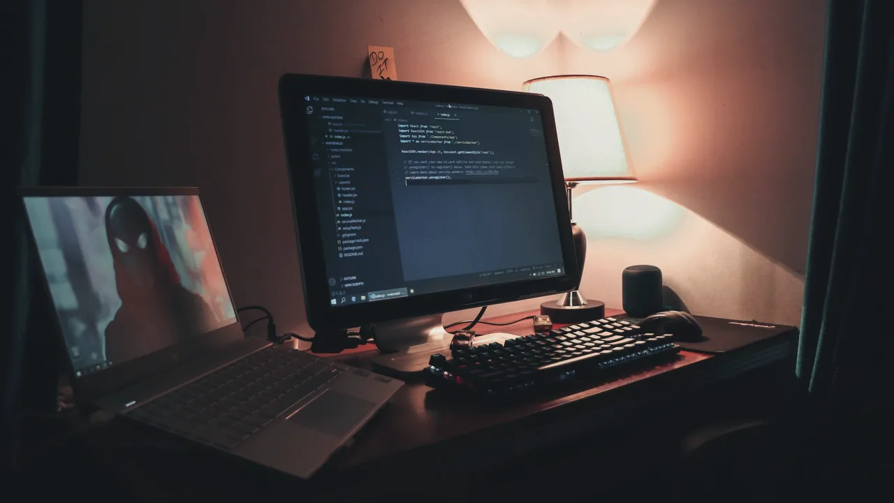 Mesa de trabalho em ambiente escuro com monitor mostrando código e luminária acesa