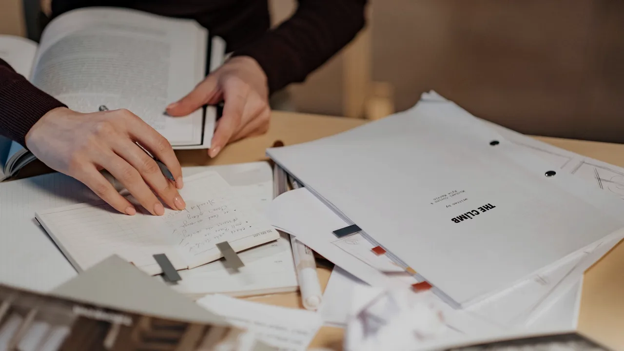 Mãos sobre uma mesa com folhas de papel e um caderno aberto, em leitura e anotação