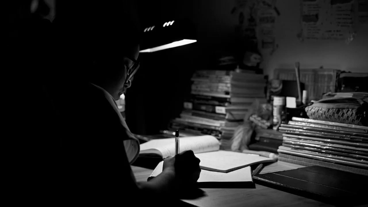 pessoa escrevendo em um caderno sob a luz de uma luminária, em ambiente escuro cercado de livros empilhados